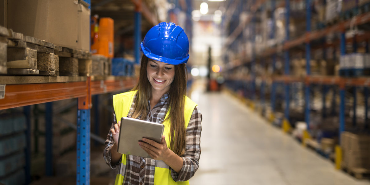 Warehouse female worker checking inventory on digital tablet in large distribution warehouse storage area.